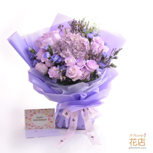 一束由18枝粉紫丁香玫瑰、淺紫色繡球、青蓮色康乃馨及紫色臘梅組成的精緻花束，用精美包裝紙和絲帶包裝，背景為柔和的室內燈光。